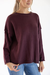 Image of a woman wearing a maroon colored slouchy sweater with blue denim jeans standing in front of a white studio background