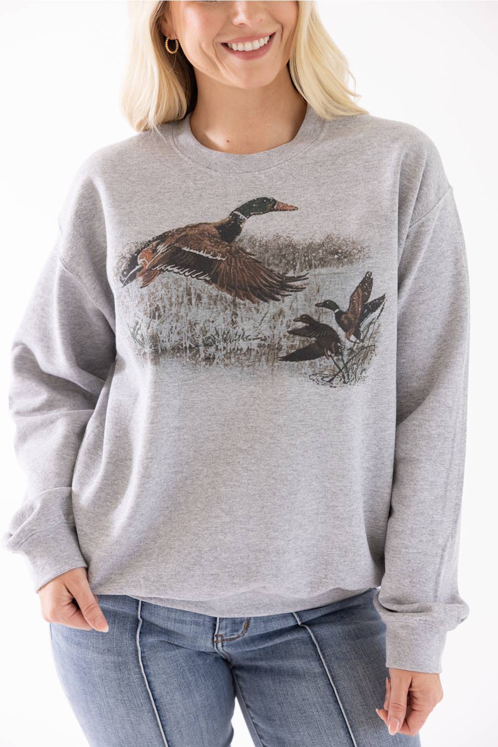 A woman wearing a light heather gray crewneck sweatshirt with a graphic print of mallards on the front, paired with blue jeans against a plain white background.