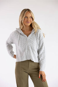 A woman modeling a light gray cable-knit zip-up hoodie with a drawstring hood, paired with olive green pants, against a plain white studio background.