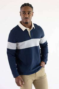 A man wearing a navy and grey color-blocked long-sleeve rugby shirt with blue jeans, standing against a plain light background.