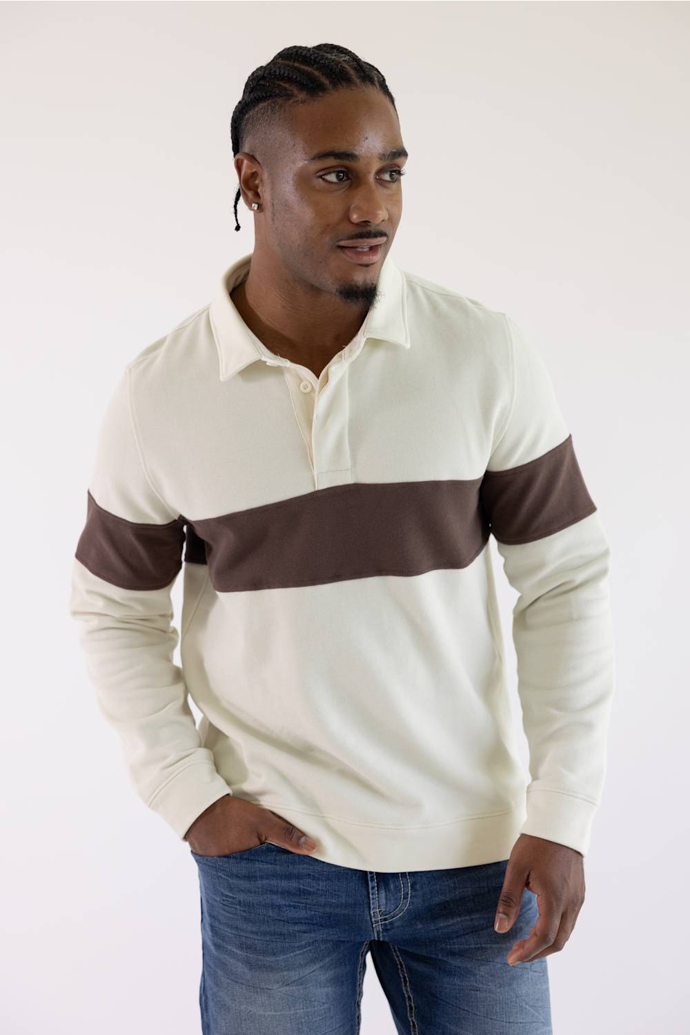 A man wearing a cream and brown color-blocked long-sleeve rugby shirt with blue jeans, standing against a plain light background.