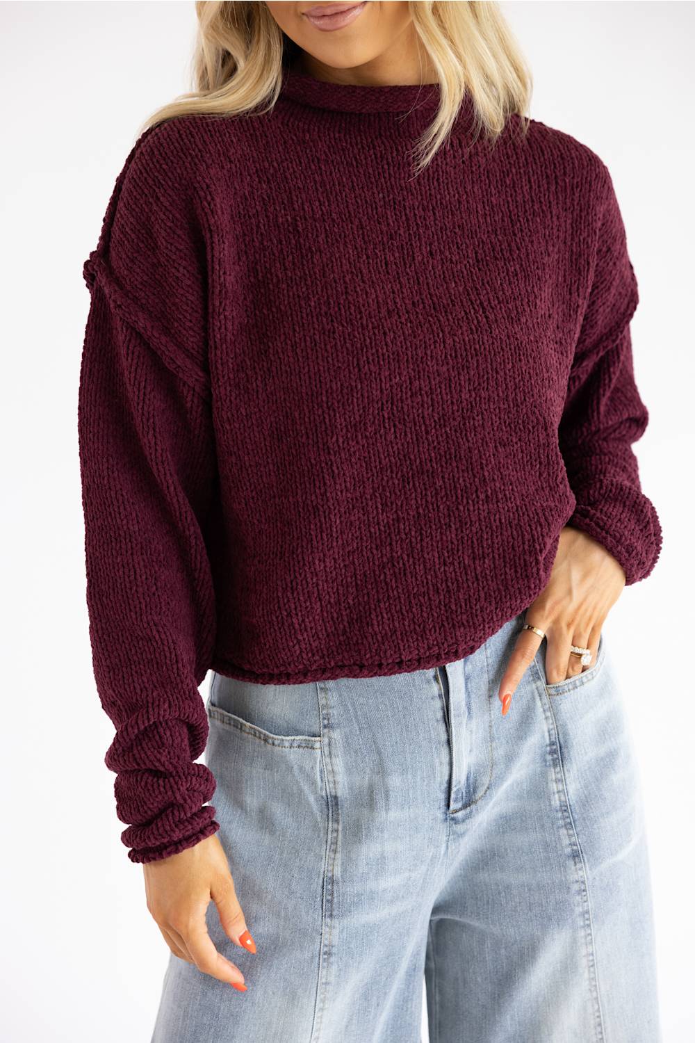 A woman wearing a ribbed knit mock neck sweater in deep burgundy, paired with light-wash blue jeans, shown from the chin down against a plain white background.