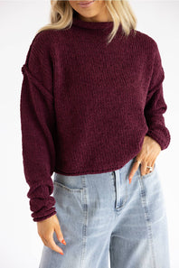 A woman wearing a ribbed knit mock neck sweater in deep burgundy, paired with light-wash blue jeans, shown from the chin down against a plain white background.