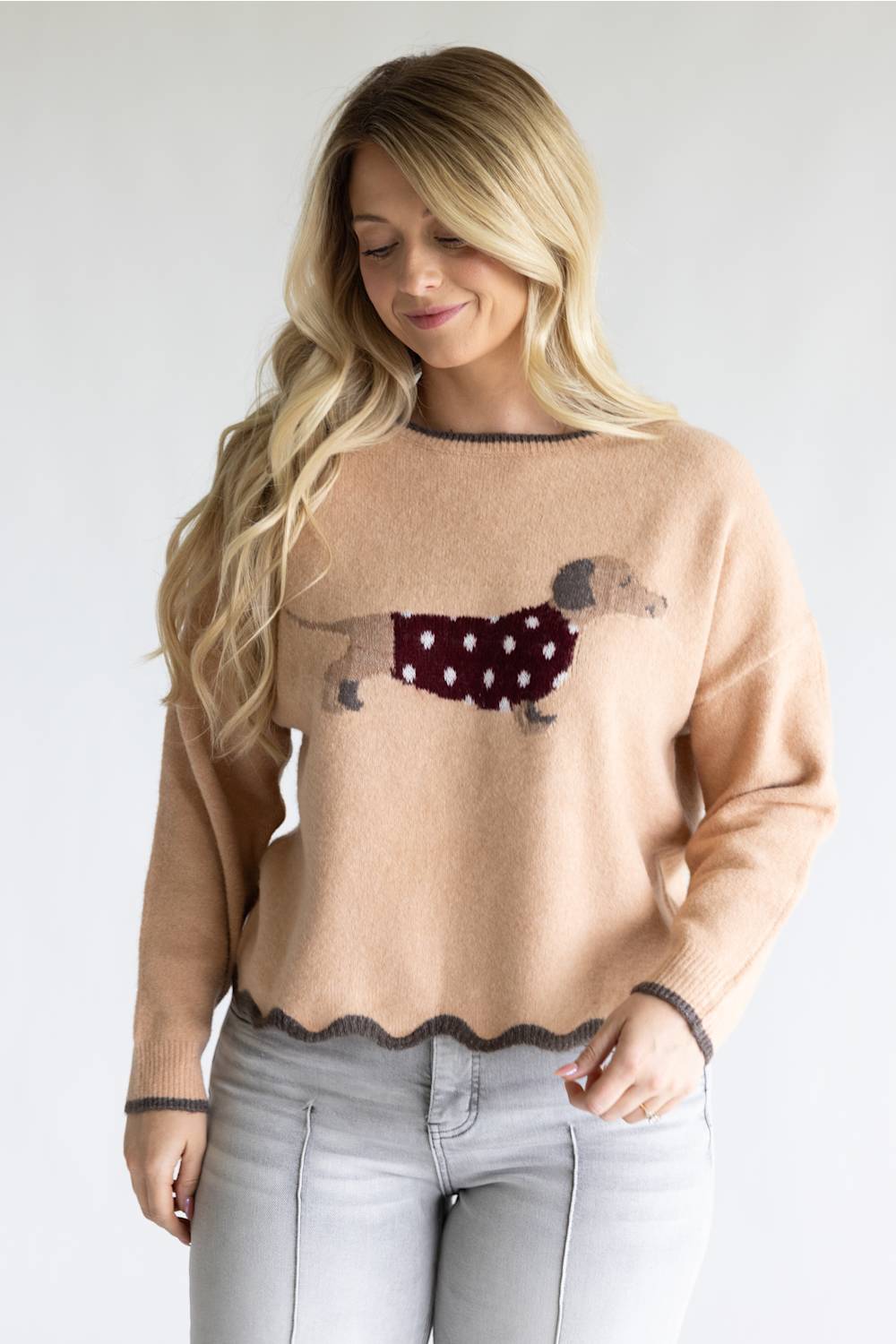 A woman wearing a beige knit sweater with a dachshund graphic wearing a burgundy polka-dot sweater, paired with light gray jeans, against a plain light gray background.