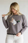 A woman wearing a gray knit sweater with a large embroidered mallard duck design on the front, paired with white ribbed pants, against a plain light gray background.