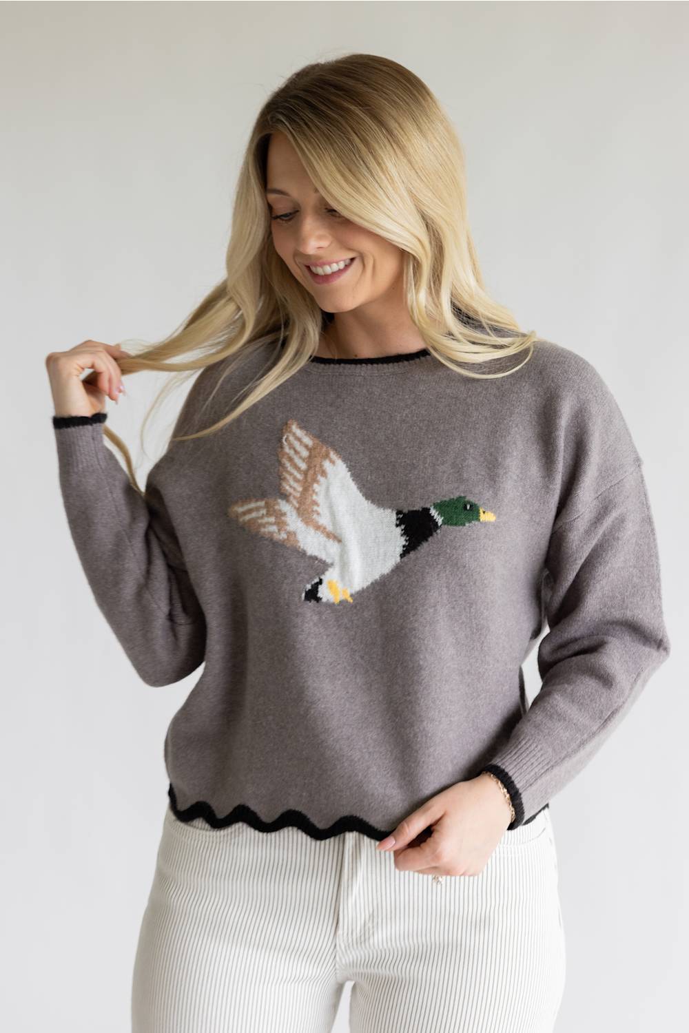 A woman wearing a gray knit sweater with a large embroidered mallard duck design on the front, paired with white ribbed pants, against a plain light gray background.