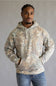 A man wearing a camouflage-patterned pullover hoodie with a kangaroo pocket and drawstrings, paired with blue jeans, standing against a neutral gray background.