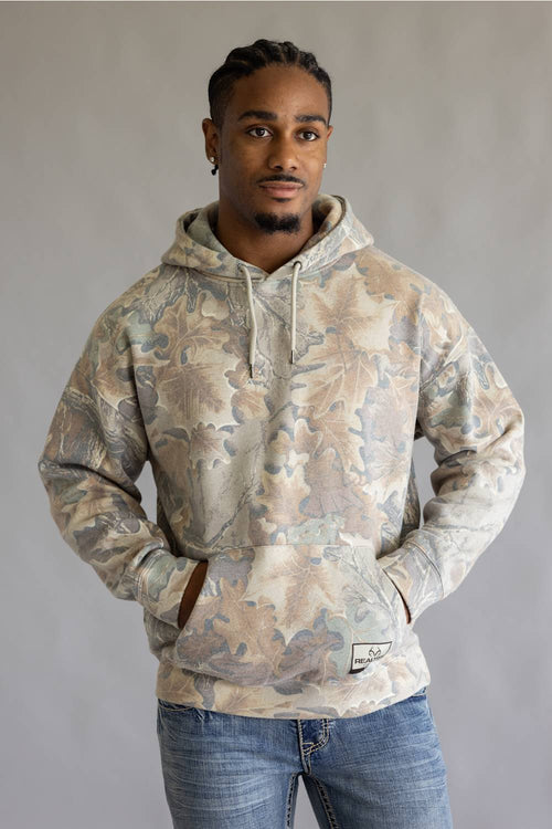 A man wearing a camouflage-patterned pullover hoodie with a kangaroo pocket and drawstrings, paired with blue jeans, standing against a neutral gray background.