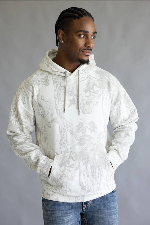 A man wearing a white and gray camo pattern hoodie with blue jeans, standing against a neutral gray studio background.