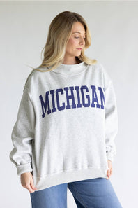 A woman wearing a light heather gray crewneck sweatshirt with 'MICHIGAN' printed in navy blue arched block letters across the chest, paired with blue jeans, against a plain light gray background.