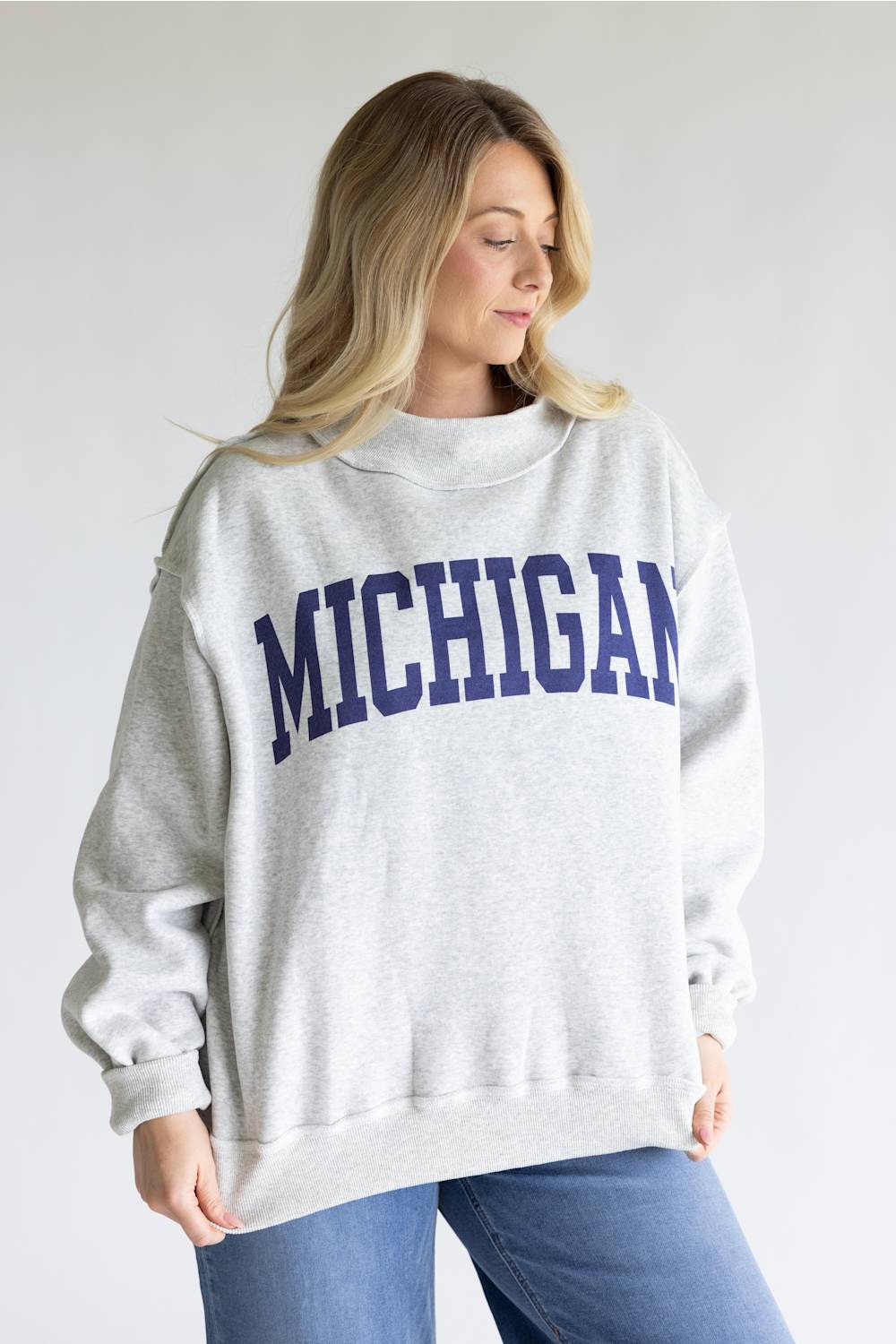 A woman wearing a light heather gray crewneck sweatshirt with 'MICHIGAN' printed in navy blue arched block letters across the chest, paired with blue jeans, against a plain light gray background.