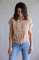 A woman wearing a beige short-sleeve cable-knit sweater and blue jeans, standing against a plain gray studio background.