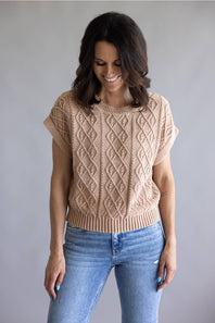 A woman wearing a beige short-sleeve cable-knit sweater and blue jeans, standing against a plain gray studio background.