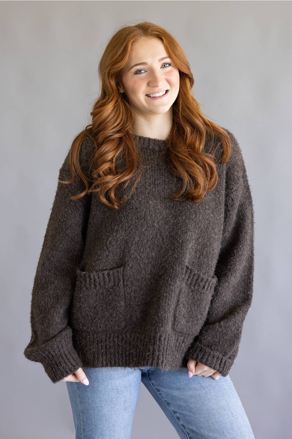 A smiling young woman with long wavy red hair wearing an oversized, chunky knit brown sweater with front pockets and light blue jeans, standing against a plain gray studio background.