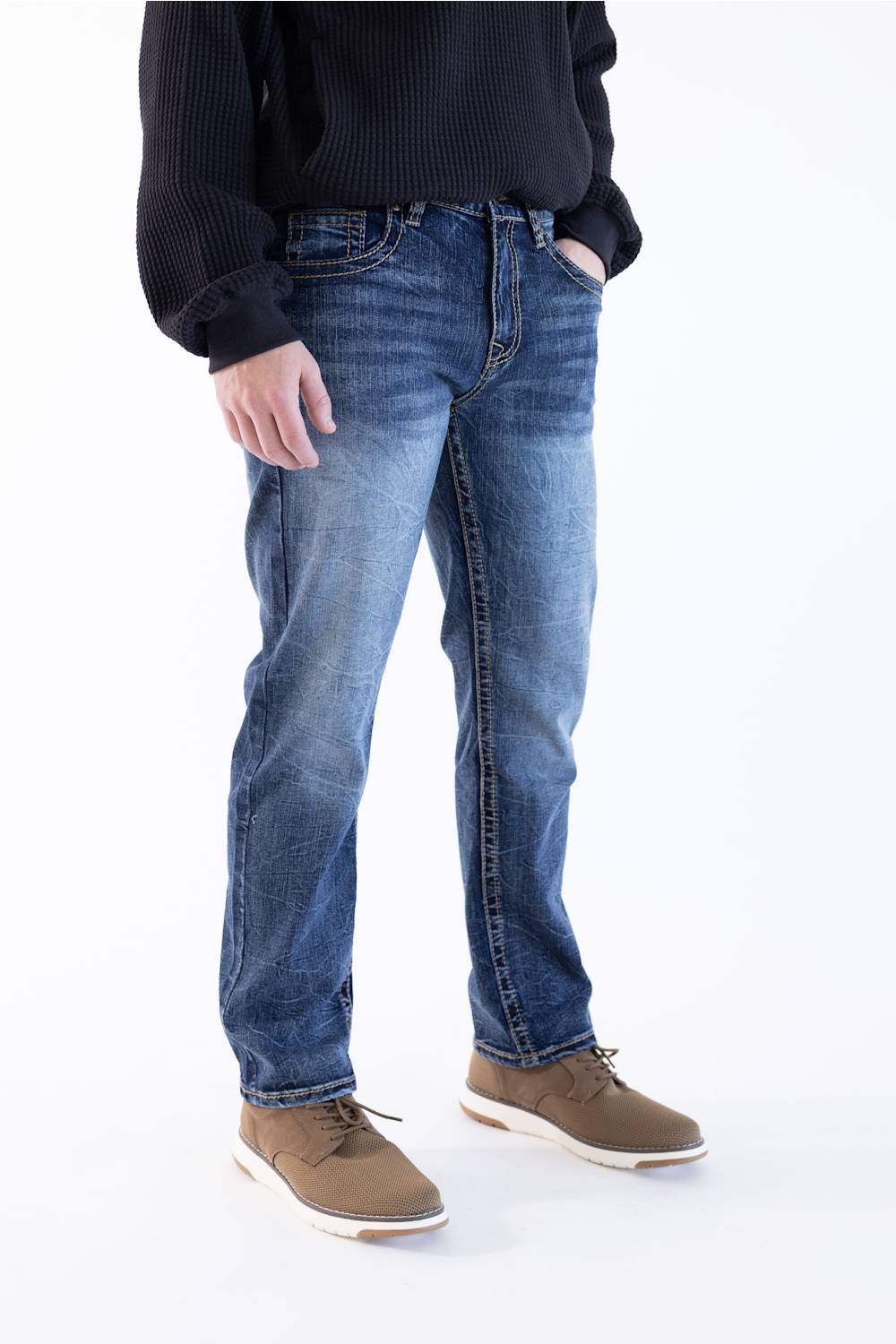 A person wearing medium-wash blue denim jeans with a black ribbed sweater and brown suede shoes, shown from the chest down against a plain white studio background.