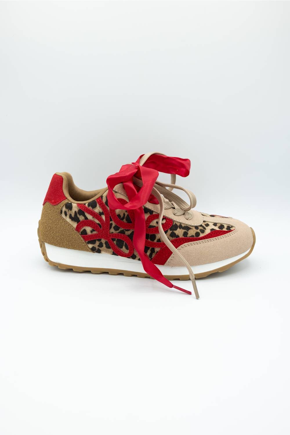 A low-top sneaker featuring a beige suede and leopard-print upper with bold red overlays, red ribbon laces, and a tan sole, shown against a plain white background.