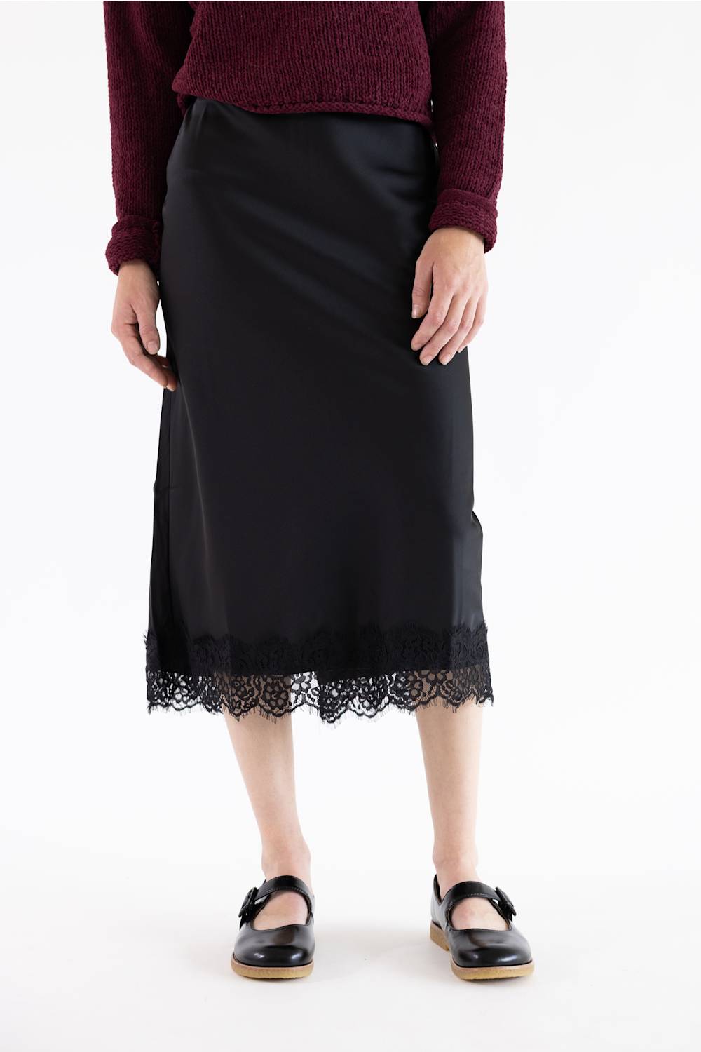 A model wearing a black midi skirt with a delicate lace hem, paired with a burgundy knit sweater and black Mary Jane shoes, shown against a plain white background.