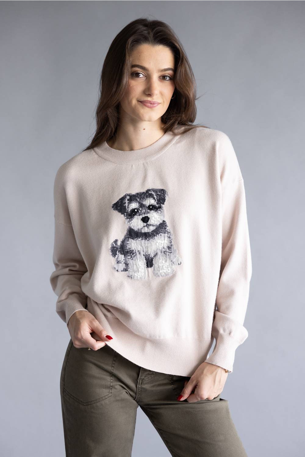 A woman wearing a light beige crewneck sweatshirt with a black and white dog graphic on the front, paired with olive green pants, against a solid gray studio background.