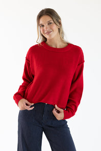 A woman wearing a red sweater with exposed seams styled with dark blue striped denim jeans standing in front of a white studio background