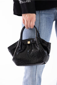 A person wearing a black knit sweater and light blue jeans holds a small black crocodile-embossed handbag with a gold-tone front clasp against a plain white background.