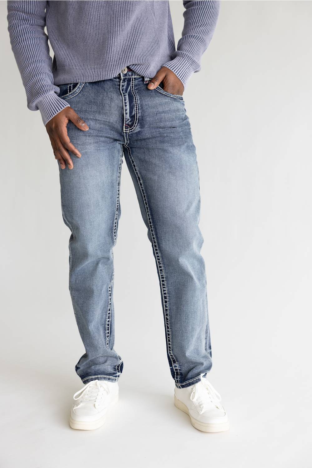 A person wearing light-wash blue denim jeans, a lavender ribbed sweater, and white sneakers, standing against a plain light gray studio background.