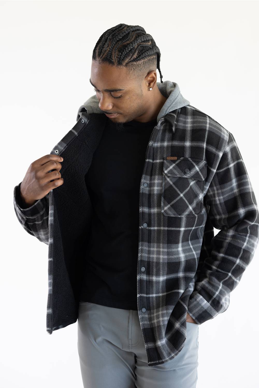 A man wearing a black and gray plaid hooded flannel shirt over a black t-shirt and light gray pants, shown against a plain white background.