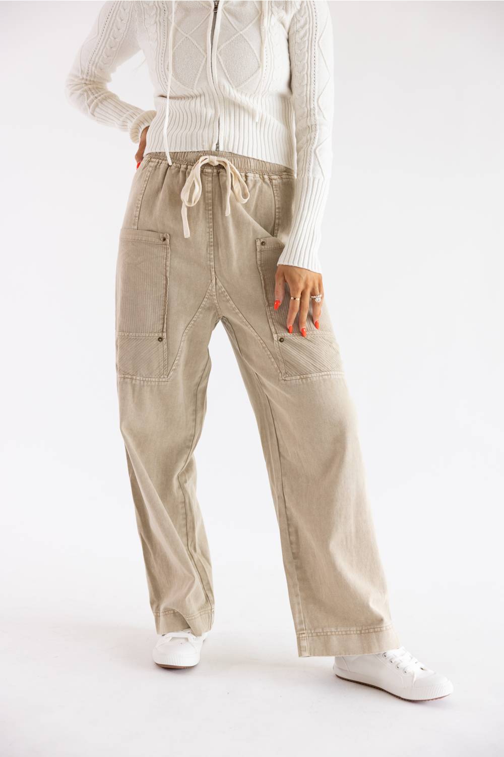 A person wearing beige wide-leg cargo-style pants with a drawstring waist, paired with a white ribbed sweater and white sneakers, photographed against a clean white studio background.
