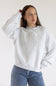 A woman wearing a light gray heather crewneck sweatshirt with embossed text reading 'SIMPLY FOR GOOD' across the chest, paired with blue jeans with small white polka dots, against a plain white background.