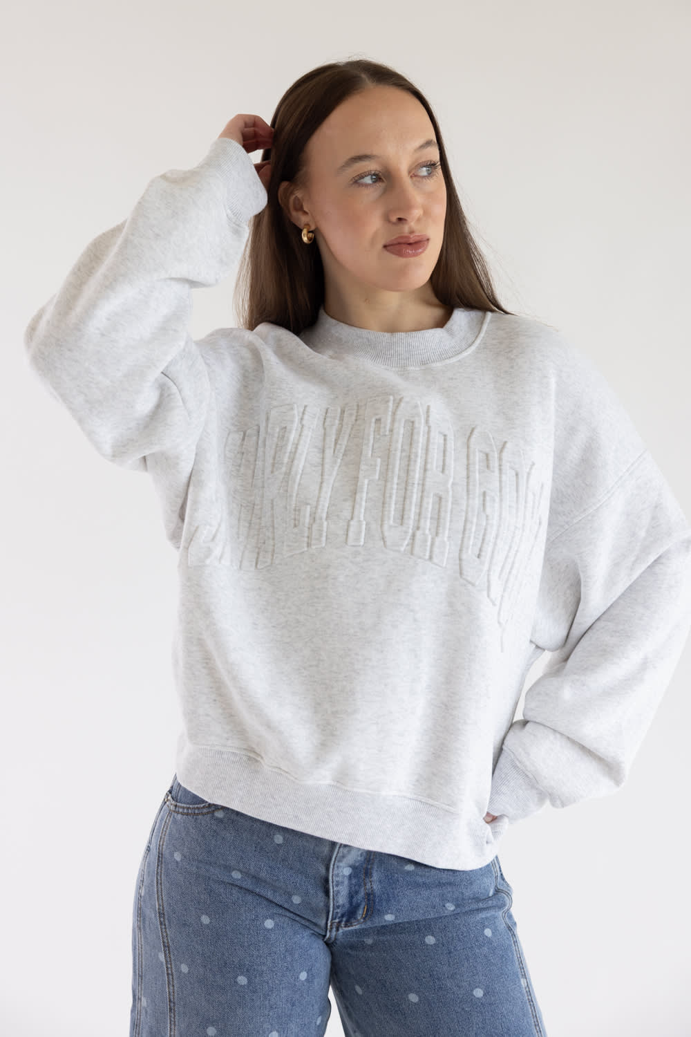 A woman wearing a light gray heather crewneck sweatshirt with embossed text reading 'SIMPLY FOR GOOD' across the chest, paired with blue jeans with small white polka dots, against a plain white background.