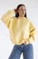 A woman wearing a pale yellow crewneck sweatshirt with embossed text reading 'Simply For Good' across the chest, paired with blue jeans with small white polka dots, against a plain white studio background.
