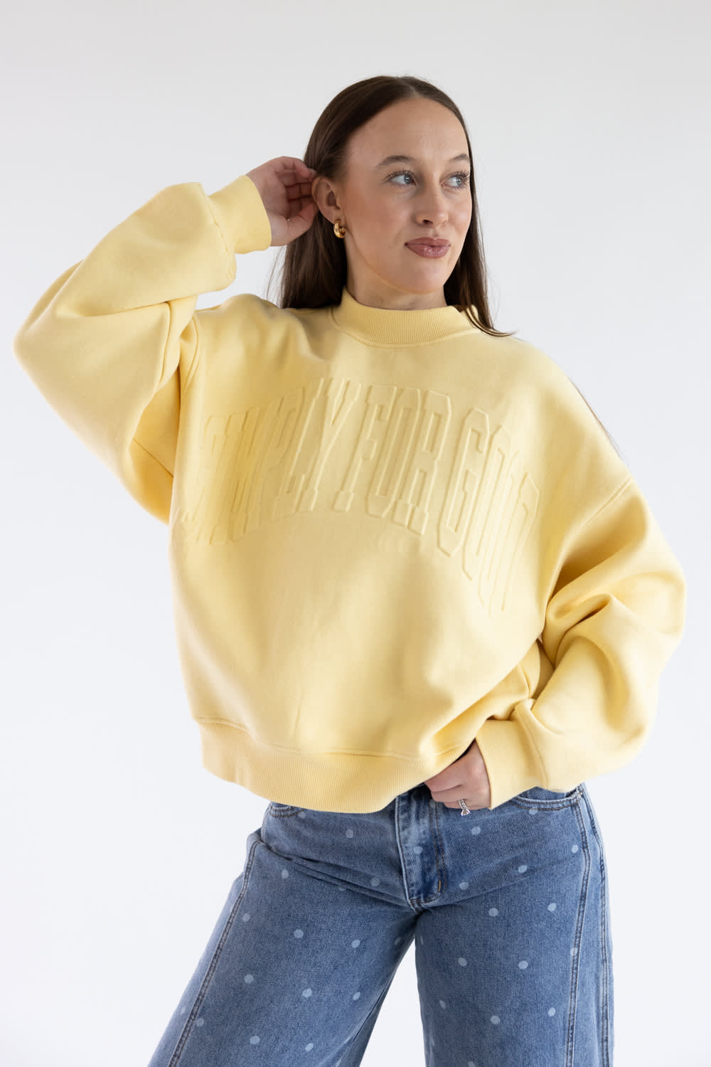 A woman wearing a pale yellow crewneck sweatshirt with embossed text reading 'Simply For Good' across the chest, paired with blue jeans with small white polka dots, against a plain white studio background.