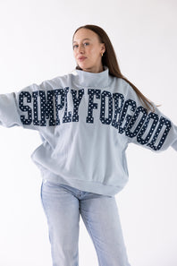A woman modeling a light blue sweatshirt with 'SIMPLY FOR GOOD' written across the back in dark navy letters with white polka dots, paired with light blue jeans and white sneakers, against a plain white background.