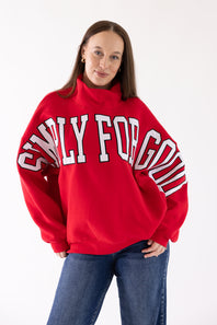 A woman wearing a bright red sweatshirt with 'SIMPLY FOR GOOD' printed in white arched letters across the chest, paired with blue jeans, against a plain white studio background.