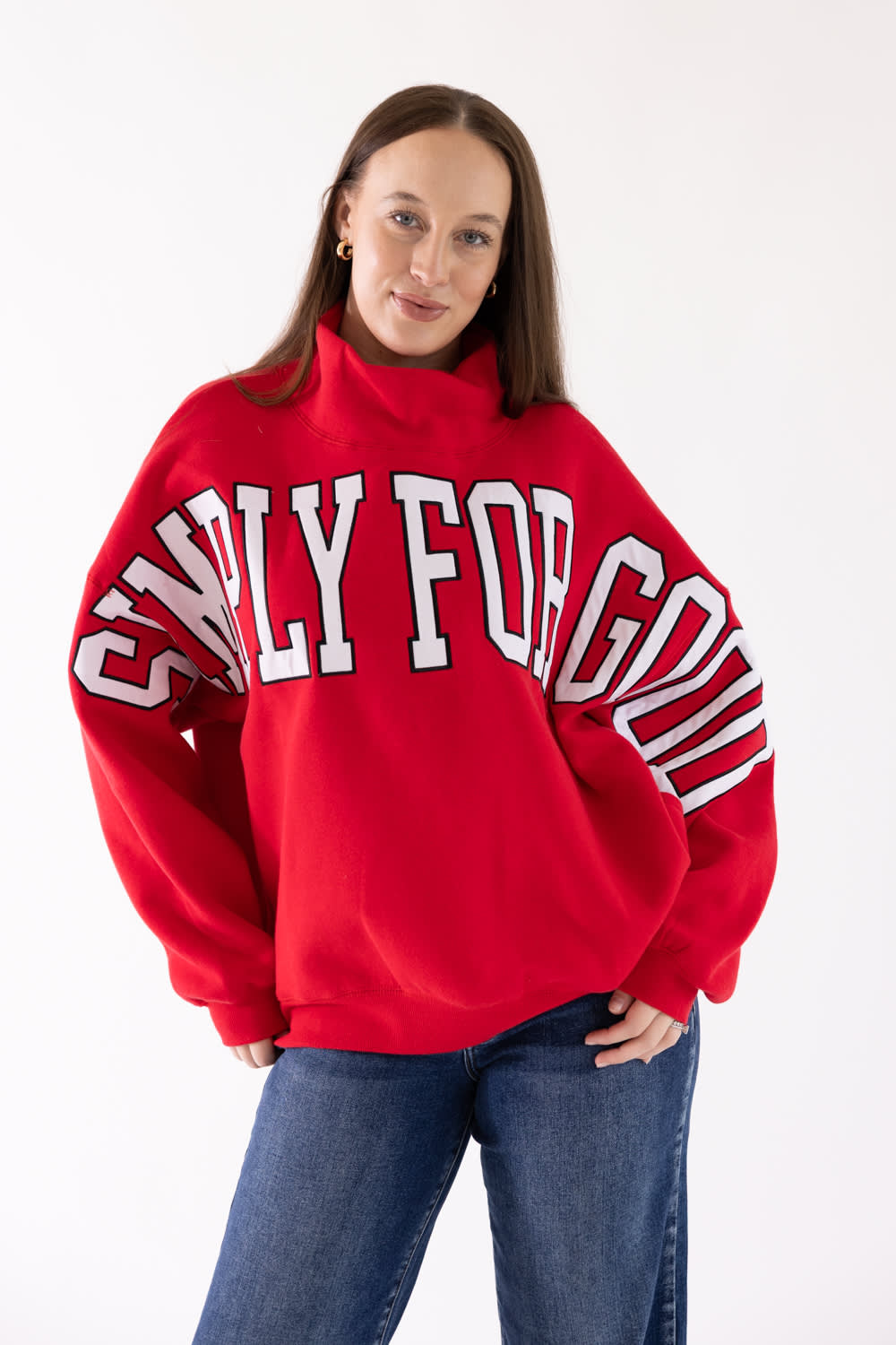 A woman wearing a bright red sweatshirt with 'SIMPLY FOR GOOD' printed in white arched letters across the chest, paired with blue jeans, against a plain white studio background.