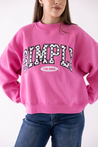 A woman wearing a bright pink crewneck sweatshirt with 'SIMPLY FOR GOOD' printed in a black-and-white polka dot arched font across the chest, paired with blue jeans.