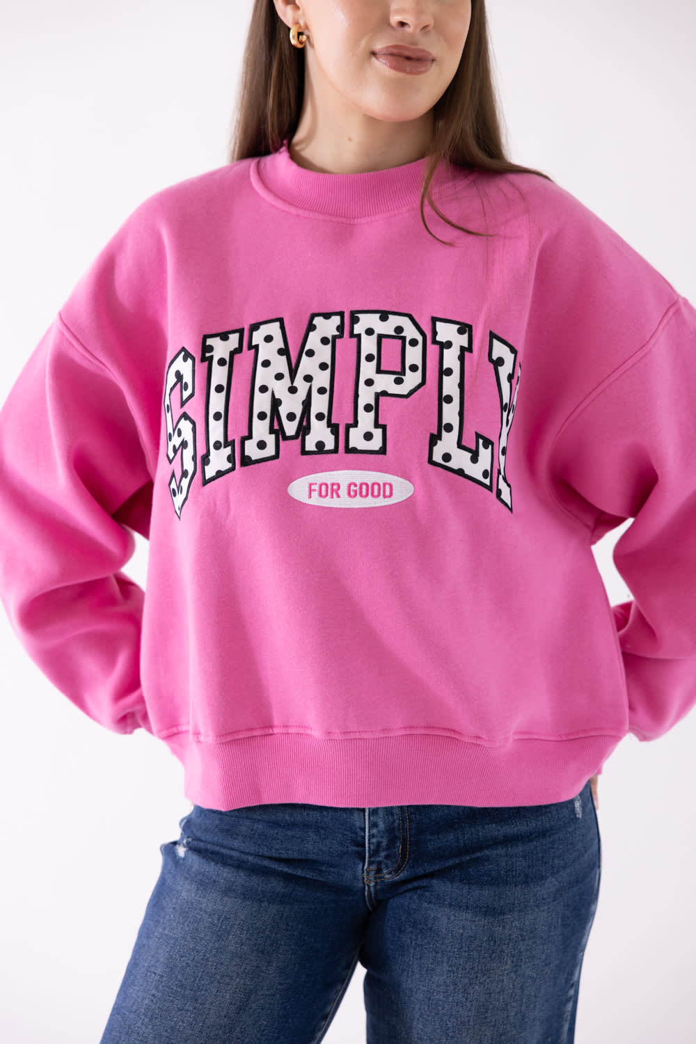 A woman wearing a bright pink crewneck sweatshirt with 'SIMPLY FOR GOOD' printed in a black-and-white polka dot arched font across the chest, paired with blue jeans.