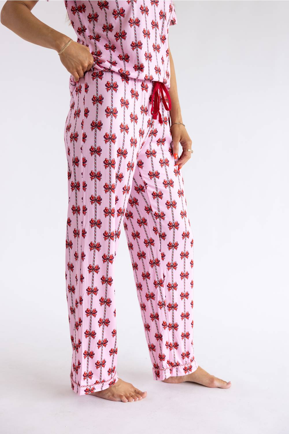 A person wearing pink pajama pants with a red bow pattern and a matching top, standing barefoot against a plain white studio background.