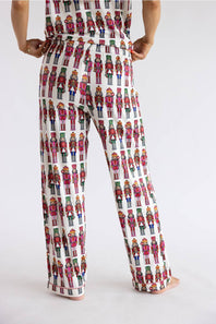 A person shown from the back wearing white wide-leg pants covered in a repeating colorful nutcracker soldier print, against a plain light gray studio background.