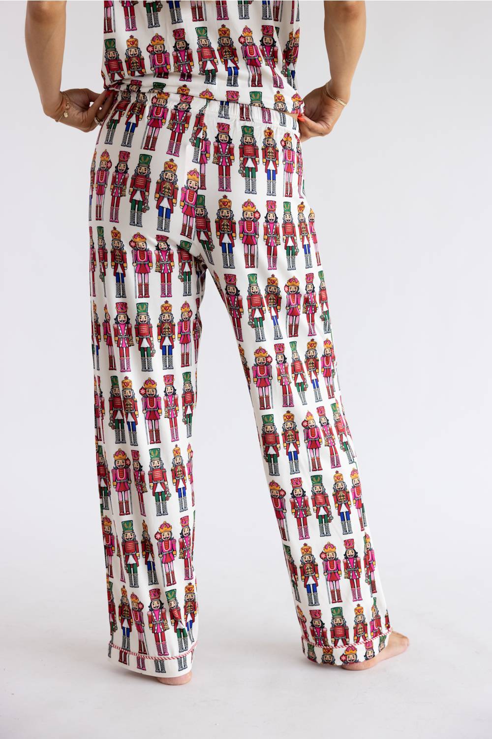 A person shown from the back wearing white wide-leg pants covered in a repeating colorful nutcracker soldier print, against a plain light gray studio background.