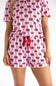 A woman wearing a pink short-sleeve romper with a repeating red bow and black line pattern, featuring a red drawstring waist, shown against a plain white background.