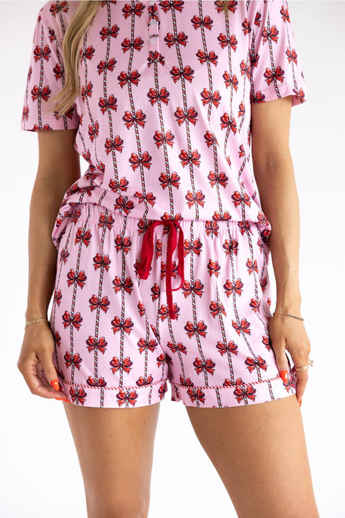 A woman wearing a pink short-sleeve romper with a repeating red bow and black line pattern, featuring a red drawstring waist, shown against a plain white background.