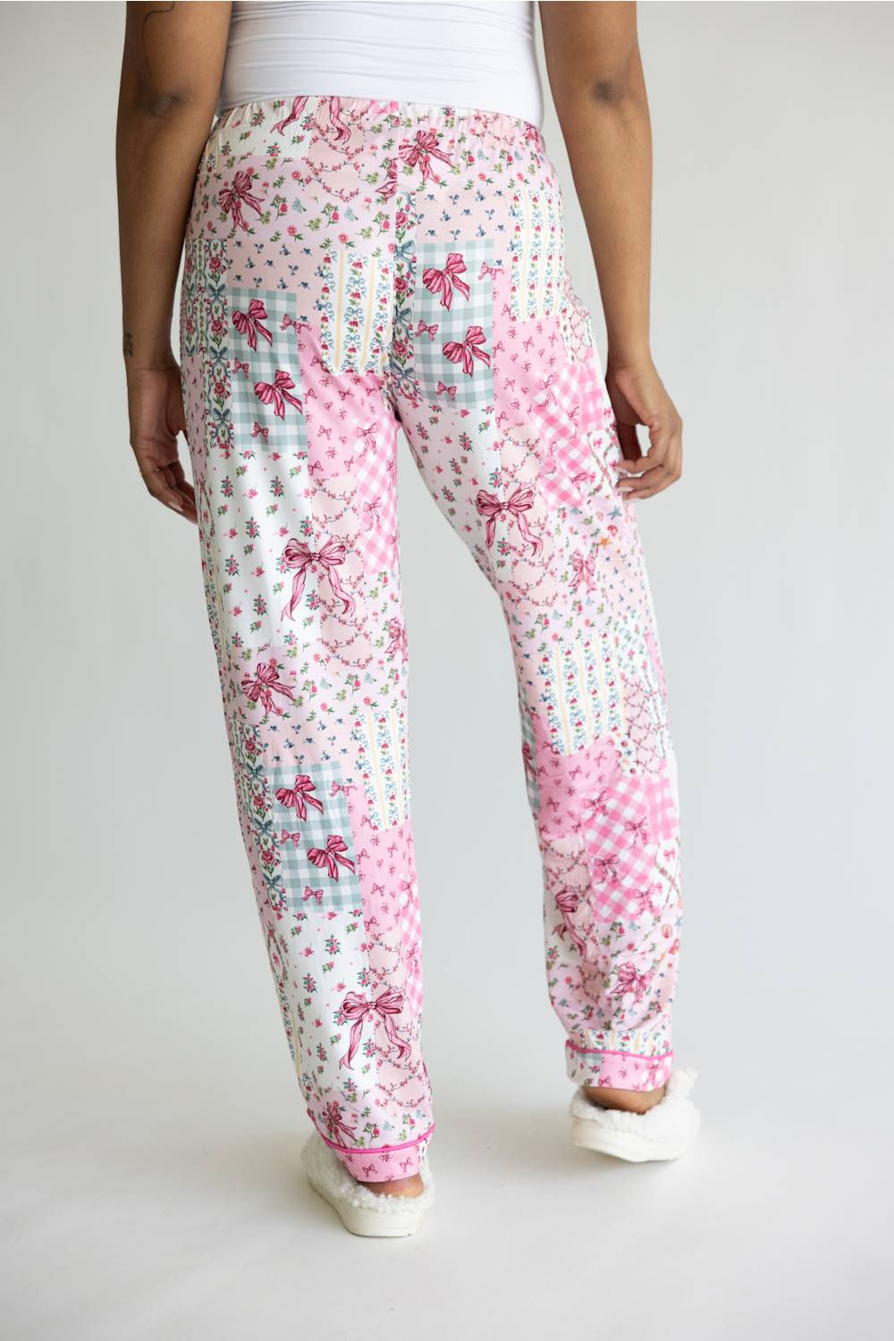 A person shown from the back wearing patchwork-patterned jogger pants with pink bows, gingham, and floral prints on a white base, paired with white slip-on shoes against a plain light gray studio background.