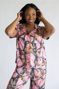 A woman modeling a matching short-sleeve pajama set with a pink camouflage tree-bark print and orange piping, against a plain light gray background.