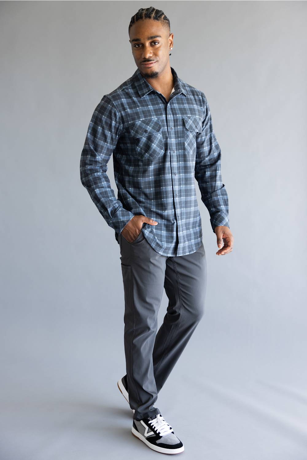A man modeling a long-sleeve plaid button-up shirt in blue and gray, paired with dark gray trousers and black and white sneakers, standing against a light gray studio background.