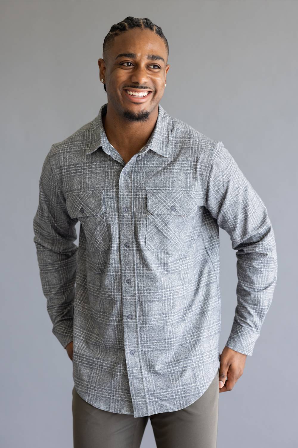 A smiling man wearing a light gray and white plaid long-sleeve button-down shirt, paired with gray pants, standing against a solid gray studio background.