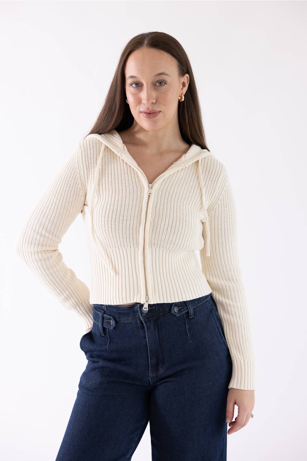 A woman modeling a cream ribbed zip-up hooded sweater with drawstrings, paired with dark blue jeans, against a plain white studio background.