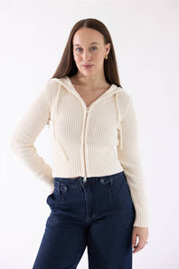 A woman modeling a cream ribbed zip-up hooded sweater with drawstrings, paired with dark blue jeans, against a plain white studio background.