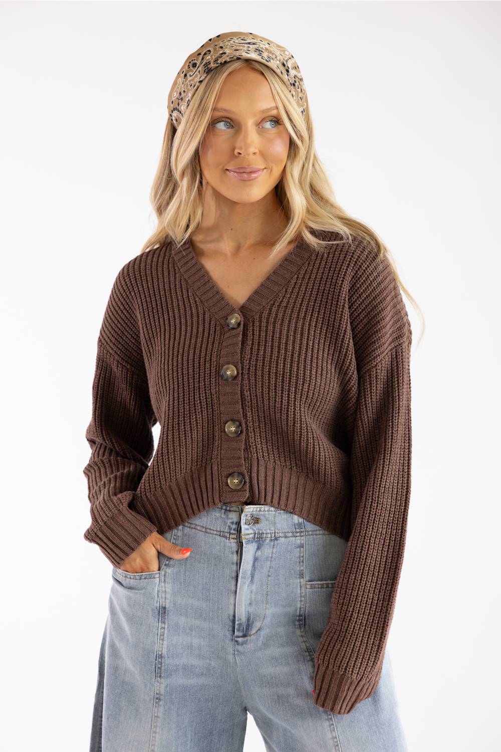A woman with wavy blonde hair wearing a patterned headband, a dark brown V-neck button-down cardigan, and light-wash denim jeans.