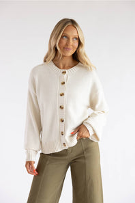 A blonde woman wearing a cream-colored knit cardigan with brown buttons and olive green pants, standing against a plain white studio background.