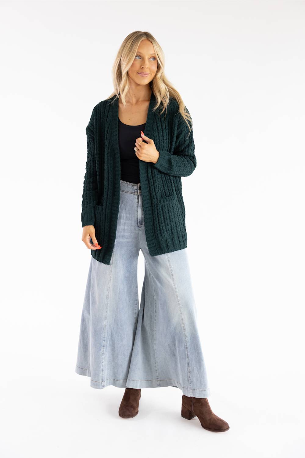A woman modeling a dark green ribbed knit cardigan over a black top and wide-leg light blue jeans, paired with brown ankle boots, against a plain white studio background.